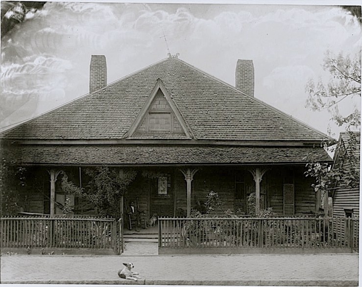 House with picket fence, man and dog seated on the porch.